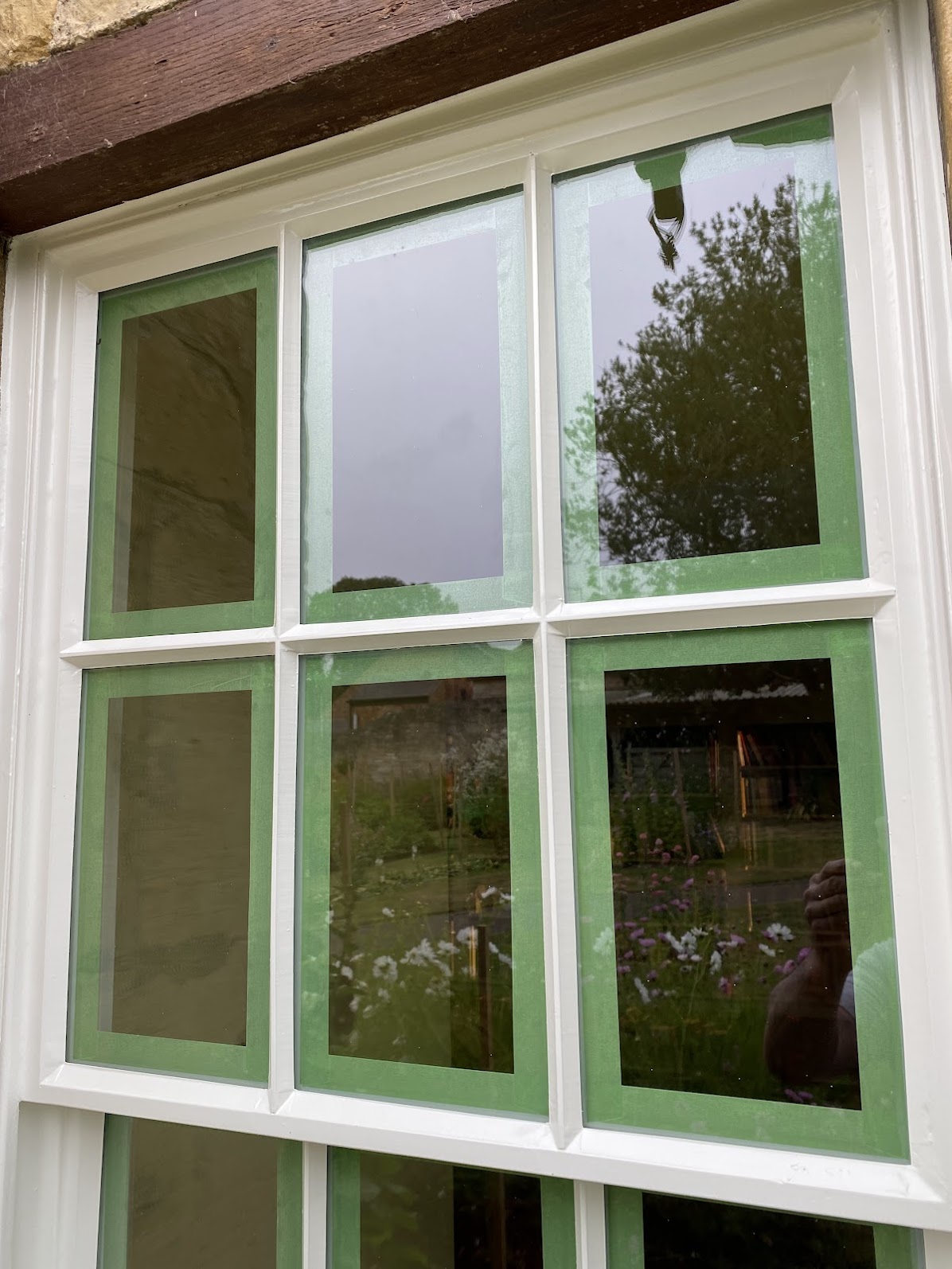 A sash window with nine panes, each bordered with green masking tape, reflecting an outdoor garden scene—ideal inspiration for Sash Window Repair South London.